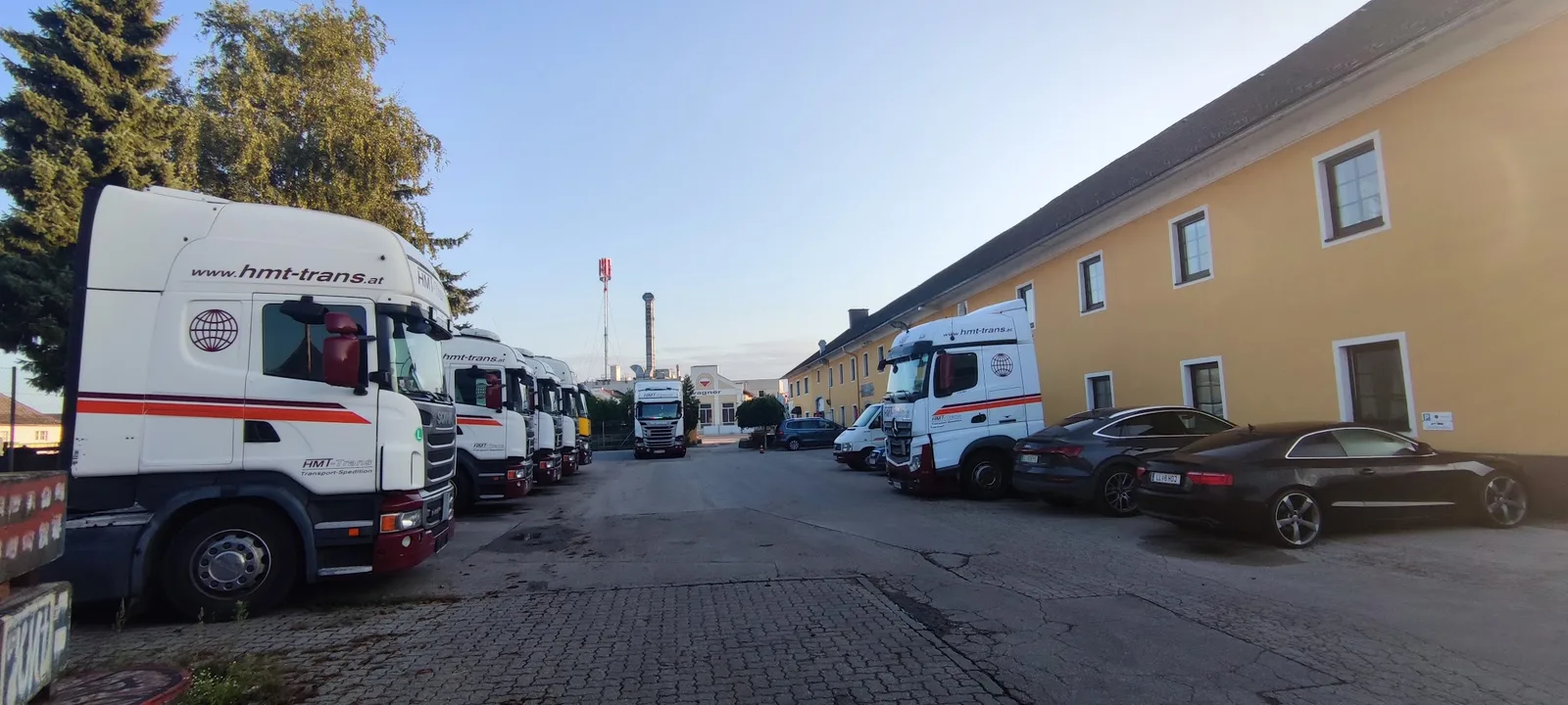 Reihe weißer HMT-Sattelzüge mit der Aufschrift hmt-trans.at neben einem gelben Gebäude